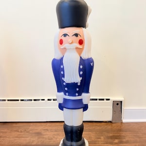 May include: A tall nutcracker figure wearing a dark blue uniform with white buttons, a black hat with a white plume, and white trousers. The figure has a white beard, moustache, and red cheeks, standing on a circular white base.