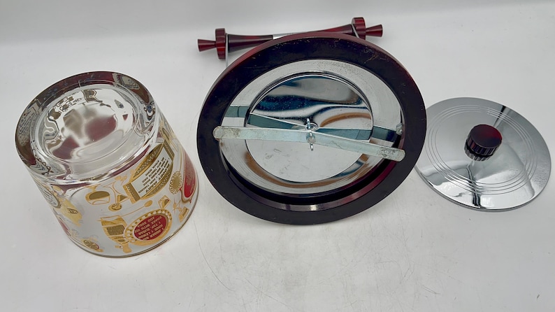 May include: An assortment of barware items, including a glass tumbler with gold and red accents, a chrome ice bucket with a dark wood rim, and a chrome lid with a red knob. The ice bucket has a chrome interior and a dark wood handle.