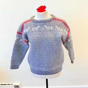 May include: A grey woollen sweater with red detailing on the shoulders and neckline. The sweater has a white geometric pattern across the chest and sleeves. The garment is displayed on a mannequin.
