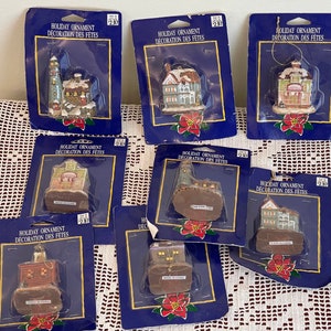 Peut inclure: Huit décorations de Noël représentant des maisons et des bâtiments miniatures. Les décorations sont emballées dans du plastique transparent avec des accents bleus et rouges. Chaque décoration est étiquetée "Holiday Ornament Decoration des Fetes" et porte une étiquette de prix de 2,97 €.