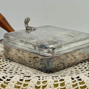 May include: A vintage silver-plated serving dish with a wooden handle and a decorative handle on the lid. The rectangular dish sits on a cream-colored, crocheted doily. The handle is a warm brown color.