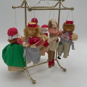 May include: Set of five handmade straw dolls hanging from a gold metal stand. The dolls are dressed in colourful fabric with red, white and green accents. The dolls are all different sizes and have different facial features.