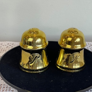 May include: A set of two gold salt and pepper shakers with a stylized letter "P" on each. The shakers are on a black tray.