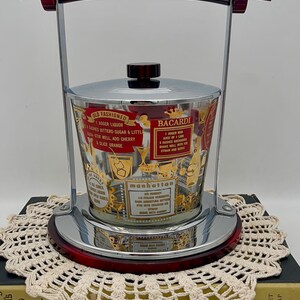 May include: Vintage cocktail ice bucket with a chrome and red frame, featuring a glass container with gold lettering and cocktail recipes. The bucket has a lid and a handle, perfect for serving drinks at a home bar.