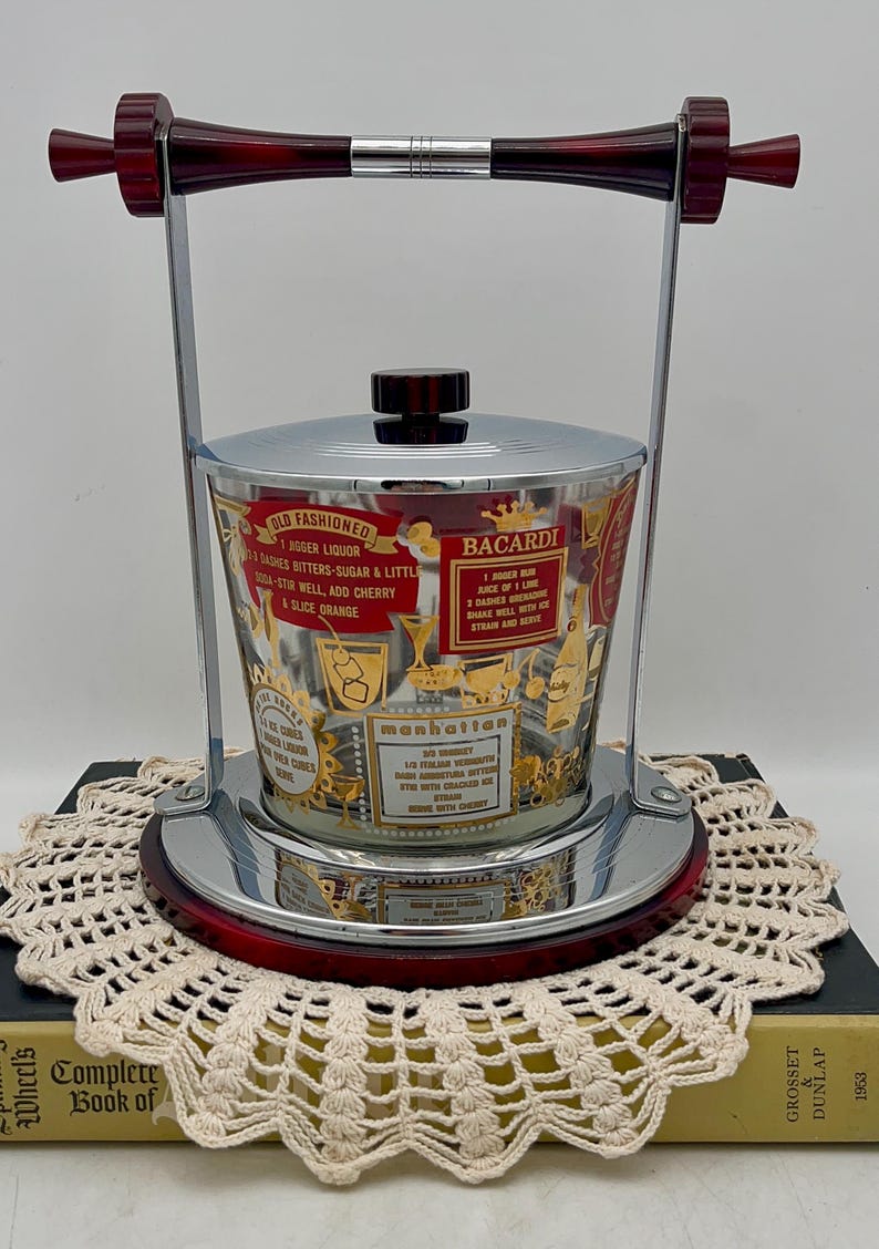 May include: Vintage cocktail ice bucket with a chrome and red frame, featuring a glass container with gold lettering and cocktail recipes. The bucket has a lid and a handle, perfect for serving drinks at a home bar.