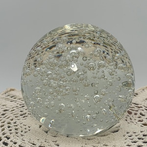 May include: A clear glass paperweight with many air bubbles inside. The spherical object rests on a cream-coloured, open-weave doily, against a plain grey background.