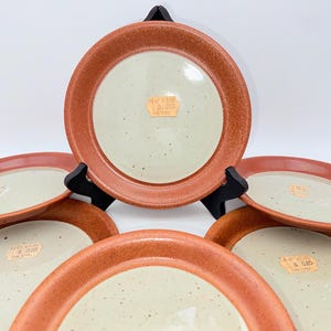 May include: A collection of six ceramic plates. Each plate features a speckled, off-white centre and a reddish-brown rim. The plates are displayed on black stands against a white backdrop. Each plate has a price tag.