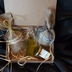 May include: A gift box filled with straw and two burlap bags tied with twine. One bag is tied with yellow twine and the other with blue twine. A clear glass bottle with a yellow liquid and a glass jar with a lid are also in the box.