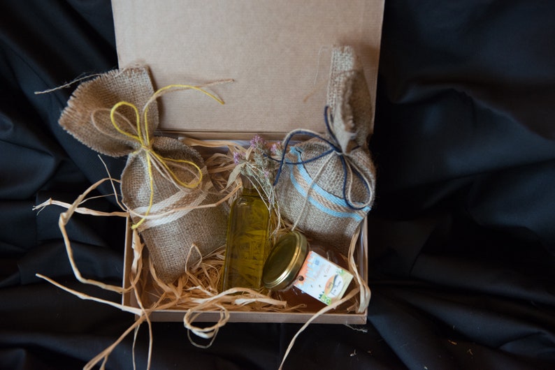 May include: A gift box filled with straw and two burlap bags tied with twine. One bag is tied with yellow twine and the other with blue twine. A clear glass bottle with a yellow liquid and a glass jar with a lid are also in the box.