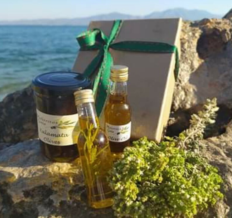 K&ouml;nnte beinhalten: Eine Geschenkbox mit gr&uuml;nem Band und Schleife, die ein Glas Kalamata-Oliven und zwei Flaschen Oliven&ouml;l enth&auml;lt. Die Flaschen sind mit "Trimino" und "Oliven&ouml;l" beschriftet.