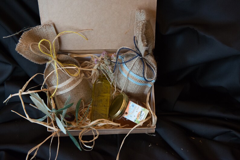 May include: A gift box filled with burlap sacks tied with twine, a glass bottle of olive oil, a jar of honey, and a sprig of olive branches. The box is lined with straw.