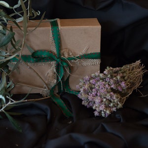 May include: A brown gift box tied with a green ribbon and burlap. The box is surrounded by green olive branches and a bundle of dried purple flowers.