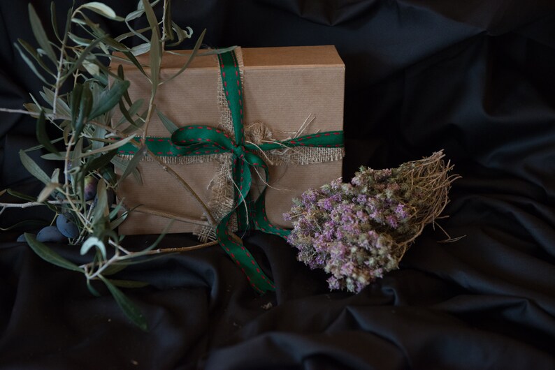 May include: A brown gift box tied with a green ribbon and burlap. The box is surrounded by green olive branches and a bundle of dried purple flowers.