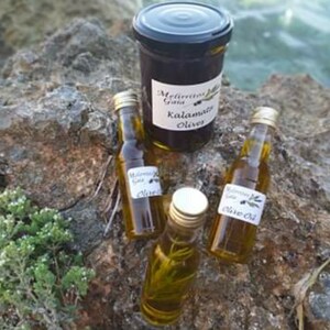 K&ouml;nnte beinhalten: Drei Glasflaschen Oliven&ouml;l mit Etiketten, auf denen "Melitini Greek Kalamata Olives Olive Oil" steht. Die Flaschen stehen auf einem Stein, hinter ihnen ein Glas mit Oliven.