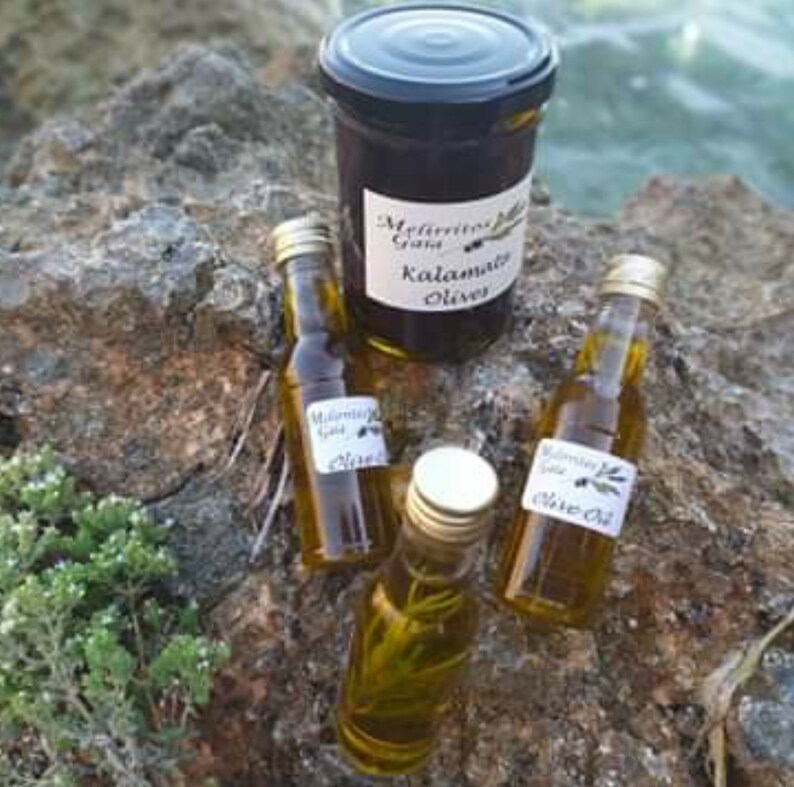 K&ouml;nnte beinhalten: Drei Glasflaschen Oliven&ouml;l mit Etiketten, auf denen "Melitini Greek Kalamata Olives Olive Oil" steht. Die Flaschen stehen auf einem Stein, hinter ihnen ein Glas mit Oliven.