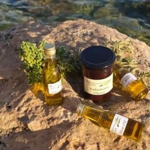 K&ouml;nnte beinhalten: Vier Flaschen Oliven&ouml;l, zwei klare Glasflaschen mit goldenen Deckeln, eine klare Glasflasche mit einem goldenen Deckel und einem gr&uuml;nen Etikett und eine dunkle Glasflasche mit einem schwarzen Deckel und einem wei&szlig;en Etikett mit der Aufschrift " Kalamata Olives". Die Flaschen stehen auf einem Stein mit gr&uuml;nen Pflanzen im Hintergrund.