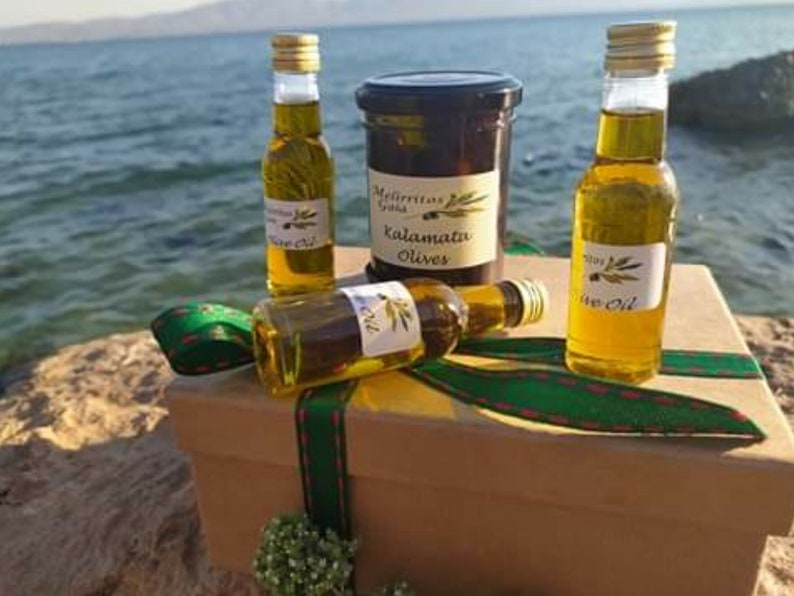 K&ouml;nnte beinhalten: Eine Geschenkbox mit einem gr&uuml;n-roten Band, die drei kleine Flaschen Oliven&ouml;l und ein Glas Kalamata-Oliven enth&auml;lt. Die Box steht auf einem Felsen am Meer.