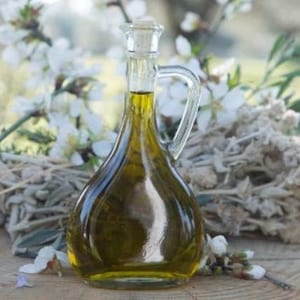 May include: A clear glass bottle with a cork stopper filled with golden olive oil. The bottle has a handle and is sitting on a bed of dried flowers.