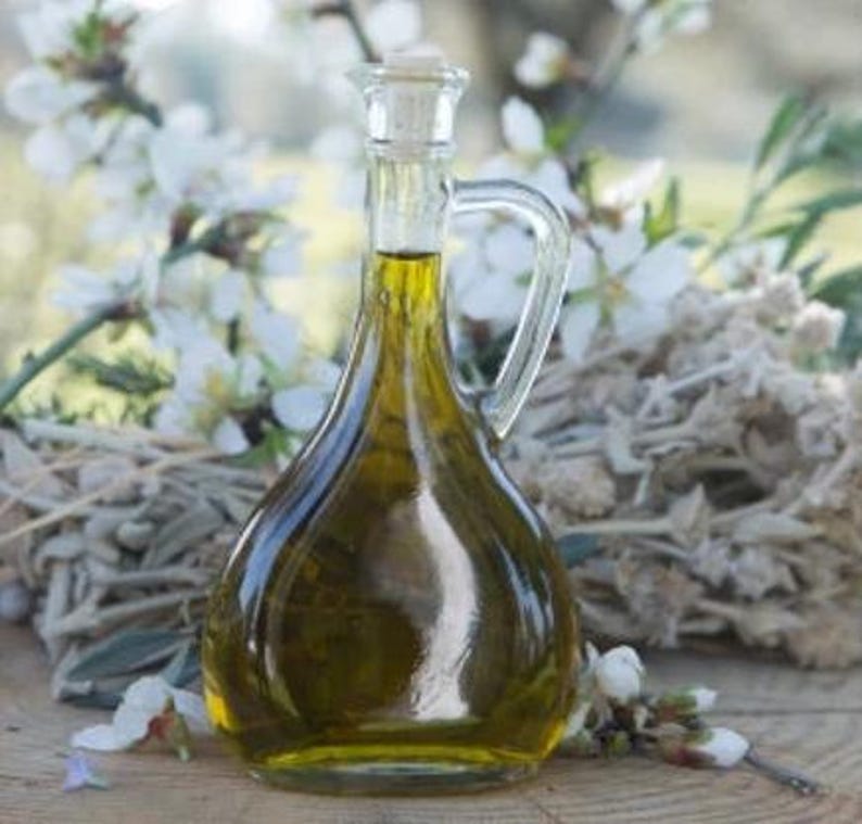 May include: A clear glass bottle with a cork stopper filled with golden olive oil. The bottle has a handle and is sitting on a bed of dried flowers.