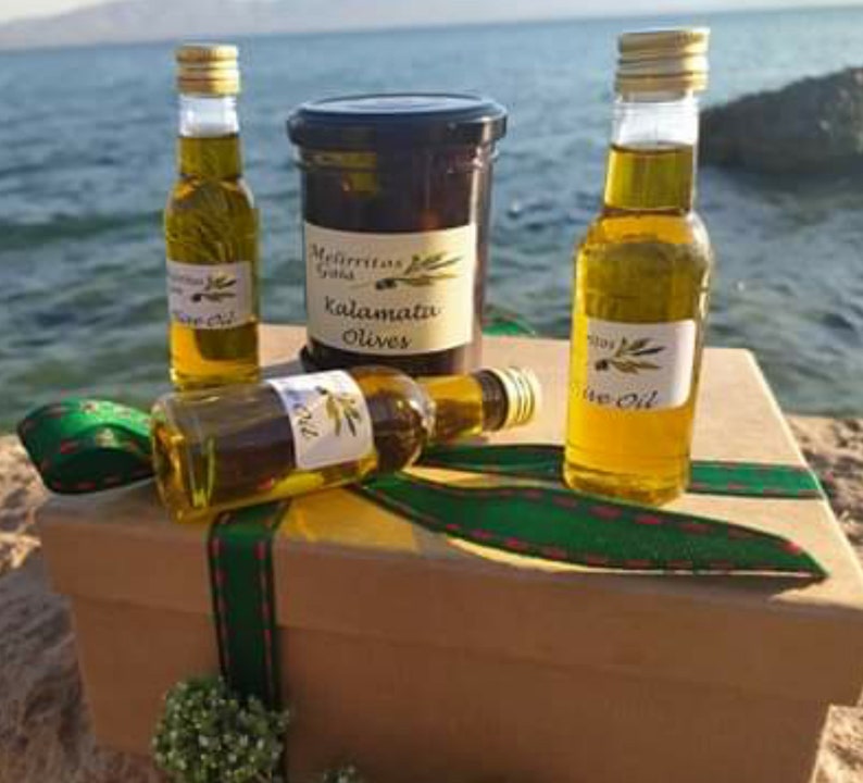 K&ouml;nnte beinhalten: Eine Geschenkbox gef&uuml;llt mit Oliven&ouml;l und Kalamata-Oliven. Die Box ist mit einem gr&uuml;n-roten Band verziert. Die Oliven&ouml;lflaschen sind aus klarem Glas mit einem gr&uuml;nen Etikett. Die Oliven befinden sich in einem Glasgef&auml;&szlig; mit einem schwarzen Etikett, auf dem "Kalamata Olives" steht.