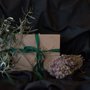 May include: A brown gift box tied with a green and red ribbon and burlap string. The box is surrounded by green olive branches and dried purple flowers.