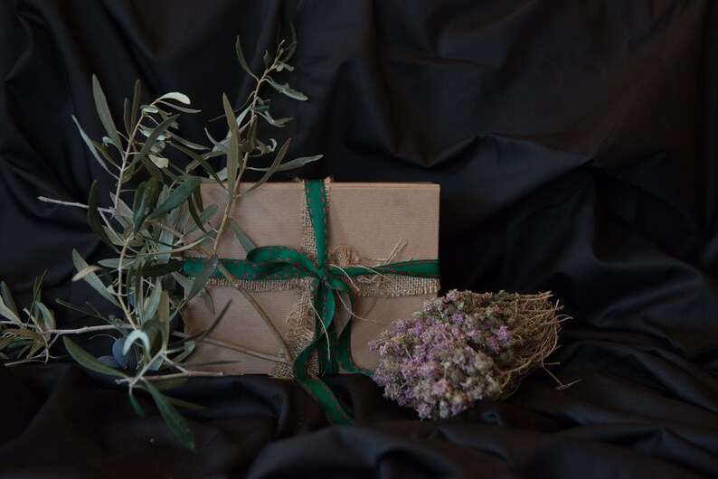 May include: A brown gift box tied with a green and red ribbon and burlap string. The box is surrounded by green olive branches and dried purple flowers.
