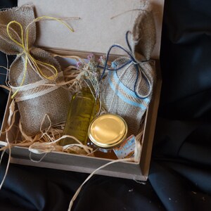 May include: A brown cardboard box filled with natural products. The box contains two small burlap sacks tied with twine, a clear glass bottle with a yellow liquid, and a glass jar with a gold lid.