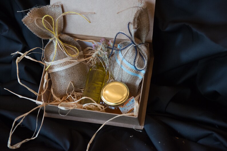 May include: A brown cardboard box filled with natural products. The box contains two small burlap sacks tied with twine, a clear glass bottle with a yellow liquid, and a glass jar with a gold lid.
