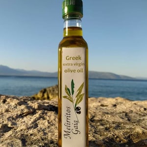 May include: A glass bottle of Greek extra virgin olive oil with a white label that says "Greek extra virgin olive oil" and "Mellirritos Gaia". The bottle is sitting on a rocky surface with a blue ocean and sky in the background.