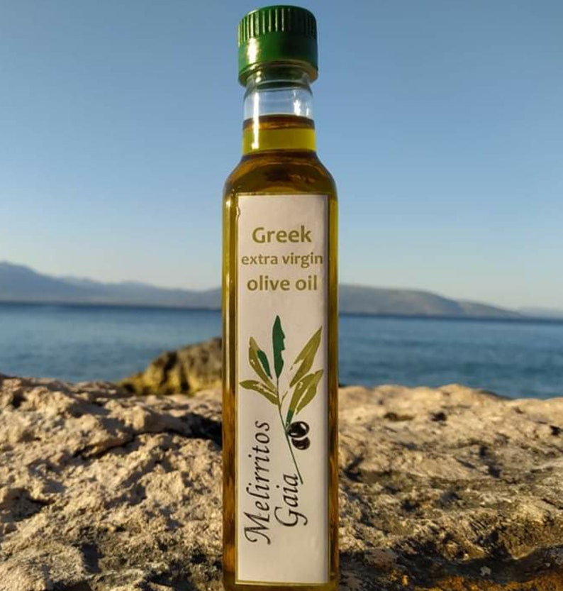 May include: A glass bottle of Greek extra virgin olive oil with a white label that says "Greek extra virgin olive oil" and "Mellirritos Gaia". The bottle is sitting on a rocky surface with a blue ocean and sky in the background.