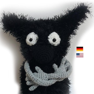 Könnte beinhalten: Ein schwarzes, gehäkeltes Plüschtier mit großen weißen Augen und grauen, gehäkelten Armen. Das Spielzeug sitzt mit gekreuzten Armen.