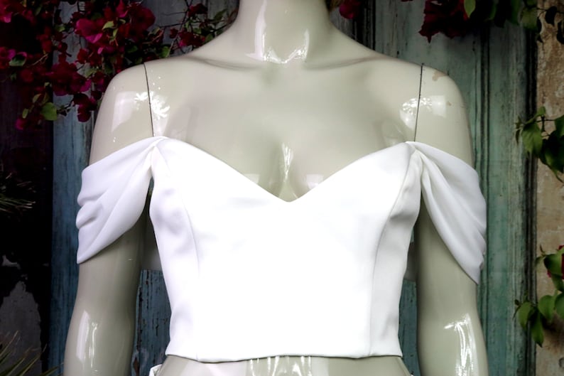 May include: A solid white cropped top featuring a sweetheart neckline and off-the-shoulder draped sleeves. The garment is fitted through the bodice with a straight hemline, showcasing a smooth, matte fabric texture across the front panel.