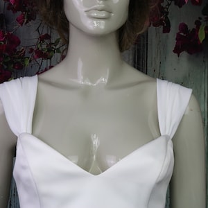 May include: A white dress featuring a sweetheart neckline and wide, gathered shoulder straps. The bodice is constructed from a smooth, solid white fabric, showcasing a clean, structured design that sits on a glossy, neutral-toned mannequin.