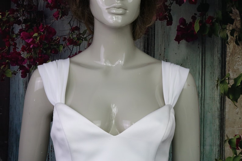 May include: A white dress featuring a sweetheart neckline and wide, gathered shoulder straps. The bodice is constructed from a smooth, solid white fabric, showcasing a clean, structured design that sits on a glossy, neutral-toned mannequin.