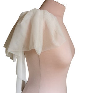 Puede incluir: Una pieza de hombro de tul blanco con un borde rizado y una larga corbata fluida. La tela es fina y delicada, y la pieza es perfecta para agregar un toque de elegancia a cualquier atuendo.