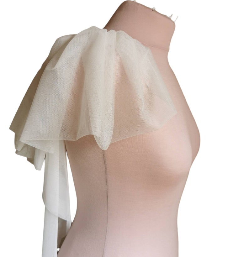 Puede incluir: Una pieza de hombro de tul blanco con un borde rizado y una larga corbata fluida. La tela es fina y delicada, y la pieza es perfecta para agregar un toque de elegancia a cualquier atuendo.