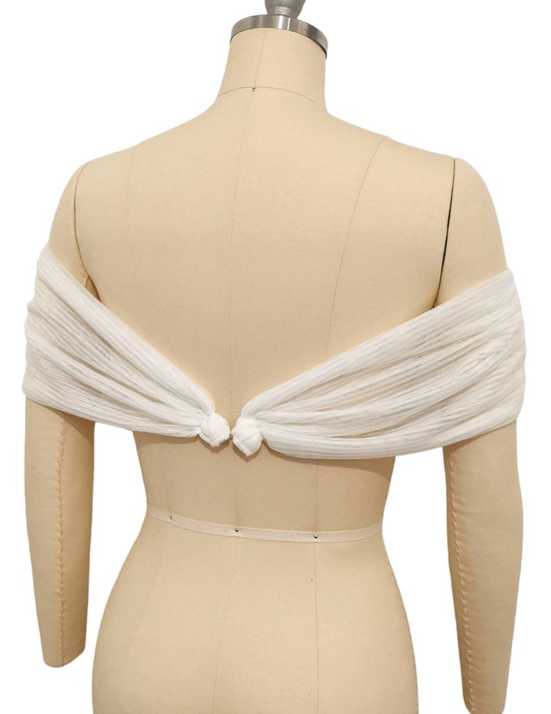 May include: White off-the-shoulder bridal shawl made of sheer fabric with a knotted detail at the back.