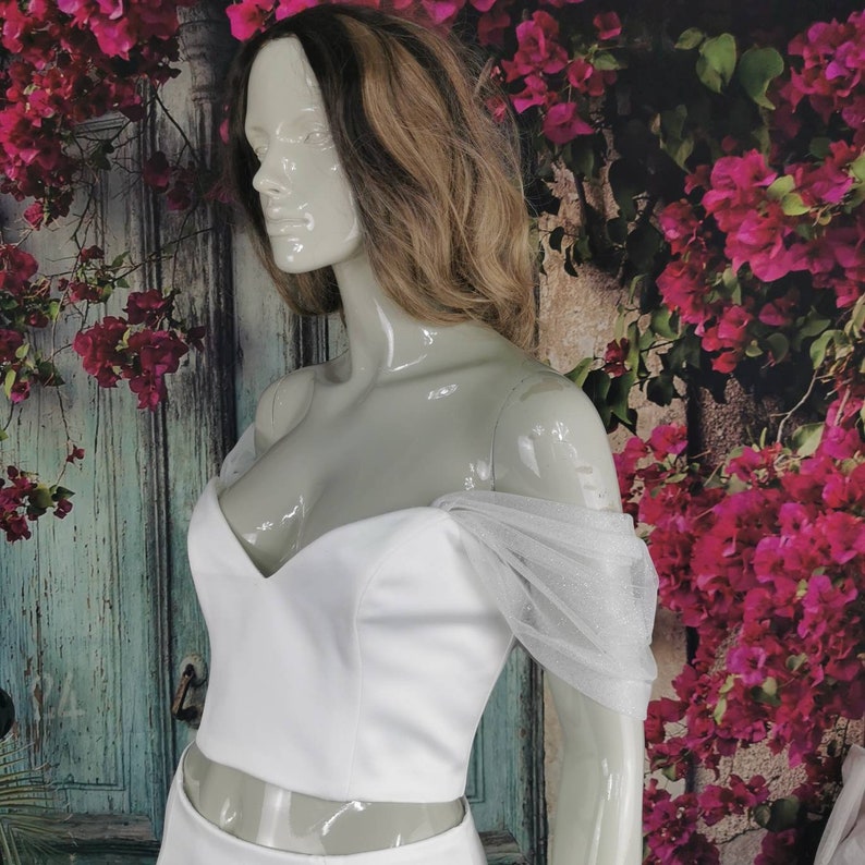 May include: A white off-the-shoulder crop top with a sweetheart neckline and tulle sleeves. The top is on a mannequin against a backdrop of pink flowers.