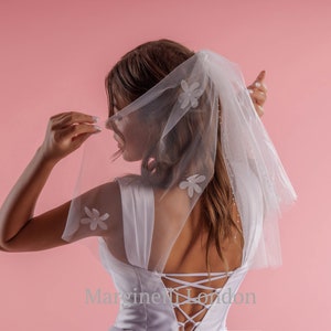 May include: A white bridal veil with delicate floral embellishments and a lace-up back detail. The veil is worn by a person with long brown hair against a pink background. The text "Margineli London" is visible at the bottom of the image.