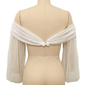 Puede incluir: Bolero de novia blanco, de hombros descubiertos, con mangas largas y transparentes. Las mangas est&aacute;n fruncidas en el codo y tienen una textura plisada. El bolero se ata en la espalda con un lazo.