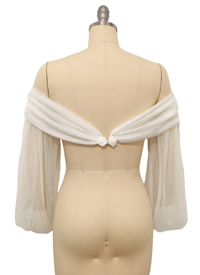 Puede incluir: Bolero de novia blanco, de hombros descubiertos, con mangas largas y transparentes. Las mangas est&aacute;n fruncidas en el codo y tienen una textura plisada. El bolero se ata en la espalda con un lazo.
