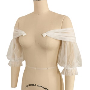 May include: White off-the-shoulder tulle sleeves with puffed sleeves and ruffled edges. The sleeves are tied at the shoulders with a bow.