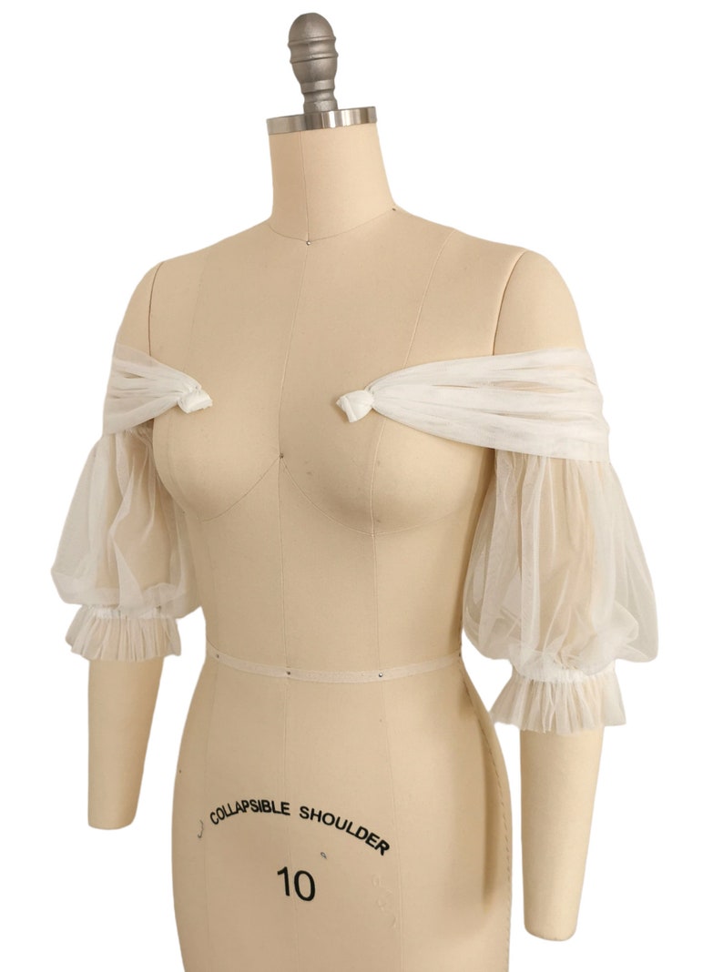 May include: White off-the-shoulder tulle sleeves with puffed sleeves and ruffled edges. The sleeves are tied at the shoulders with a bow.