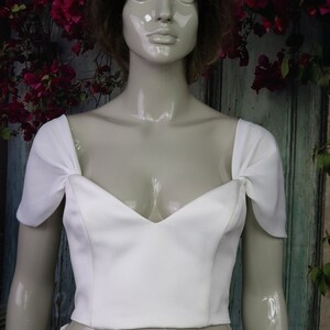 May include: A white cropped top featuring a sweetheart neckline and short, gathered cap sleeves. The garment is constructed with a fitted bodice that ends at the waistline, showcasing a clean, solid white fabric with structured seams.