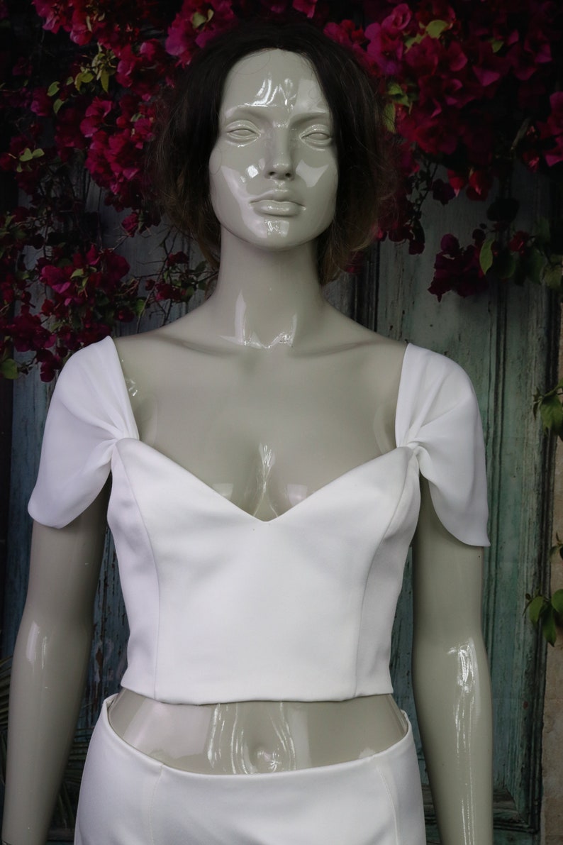 May include: A white cropped top featuring a sweetheart neckline and short, gathered cap sleeves. The garment is constructed with a fitted bodice that ends at the waistline, showcasing a clean, solid white fabric with structured seams.