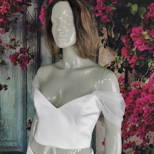 May include: White off-the-shoulder crop top with a sweetheart neckline and sheer tulle sleeves. The top is made of a smooth, satin-like fabric.