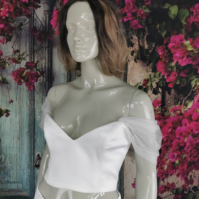 May include: White off-the-shoulder crop top with a sweetheart neckline and sheer tulle sleeves. The top is made of a smooth, satin-like fabric.