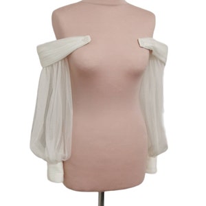 Puede incluir: Una blusa blanca de hombros descubiertos con mangas transparentes y fruncidas. La blusa se muestra en un maniqu&iacute;.