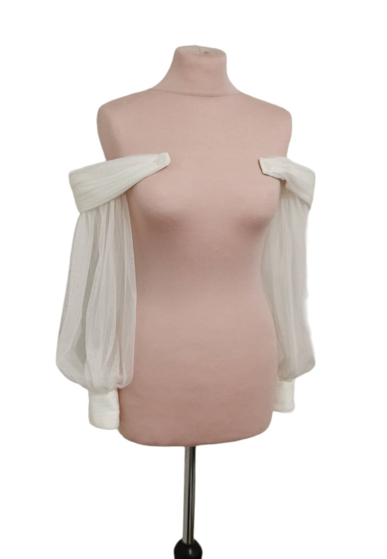 Puede incluir: Una blusa blanca de hombros descubiertos con mangas transparentes y fruncidas. La blusa se muestra en un maniqu&iacute;.
