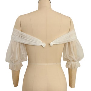May include: White off-the-shoulder tulle bridal bolero with puffed sleeves and a gathered detail at the wrist.